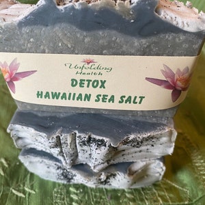 May include: Three bars of gray and white soap with black flecks. The soap is labeled "Unfolding Health DETOX HAWAIIAN SEA SALT".