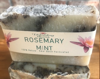 May include: Two bars of handmade soap, one on top of the other. The top bar is white with black flecks and has a label that reads "Unfolding Health, Rosemary Mint, 100% Natural Small Batch Handcrafted". The bottom bar is a similar color and texture.