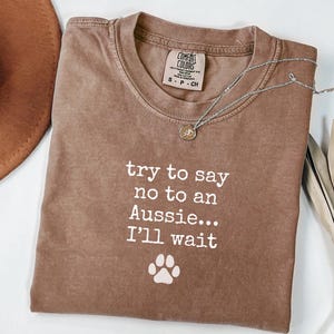 May include: A brown Comfort Colors t-shirt with the text "try to say no to an Aussie... I'll wait" and a paw print graphic. A silver necklace with a pendant is on the shirt.