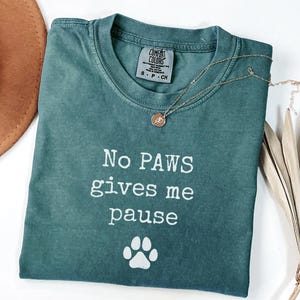 May include: A teal Comfort Colors t-shirt with the text "No PAWS gives me pause" and a paw print graphic. A gold necklace with a pendant is draped across the shirt. A brown hat and dried plant are in the background.