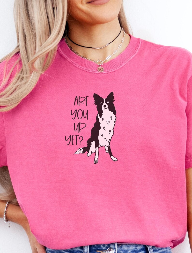 Puede incluir: Camiseta rosa brillante con una ilustraci&oacute;n en blanco y negro de un perro Border Collie. El texto "ARE YOU UP YET?" est&aacute; impreso encima del perro. La camiseta tiene mangas cortas.
