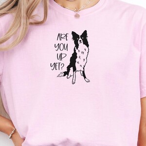 Puede incluir: Camiseta rosa claro con la frase "ARE YOU UP YET?" y una ilustraci&oacute;n en blanco y negro de un perro Border Collie. El perro est&aacute; sentado con las patas delanteras extendidas.