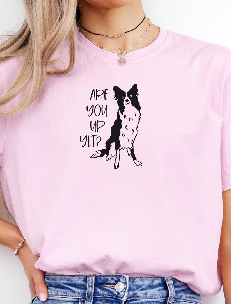 Puede incluir: Camiseta rosa claro con la frase "ARE YOU UP YET?" y una ilustraci&oacute;n en blanco y negro de un perro Border Collie. El perro est&aacute; sentado con las patas delanteras extendidas.
