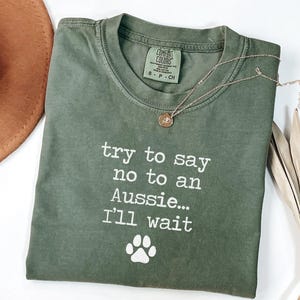 May include: Olive green t-shirt with the text "try to say no to an Aussie... I'll wait" and a paw print graphic. A gold necklace with a pendant is on the shirt. A brown hat is visible in the background.