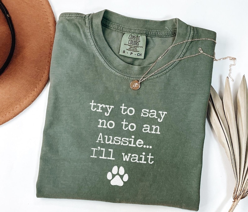 May include: Olive green t-shirt with the text "try to say no to an Aussie... I'll wait" and a paw print graphic. A gold necklace with a pendant is on the shirt. A brown hat is visible in the background.