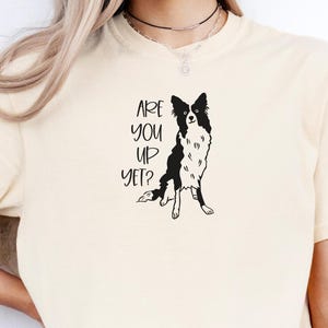 Puede incluir: Camiseta color crema con un gr&aacute;fico en blanco y negro de un perro Border Collie. El texto "ARE YOU UP YET?" est&aacute; impreso en negro encima de la ilustraci&oacute;n del perro. La camiseta tiene cuello redondo.