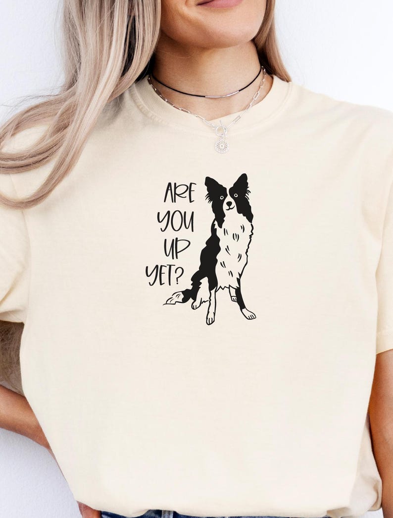 Puede incluir: Camiseta color crema con un gr&aacute;fico en blanco y negro de un perro Border Collie. El texto "ARE YOU UP YET?" est&aacute; impreso en negro encima de la ilustraci&oacute;n del perro. La camiseta tiene cuello redondo.