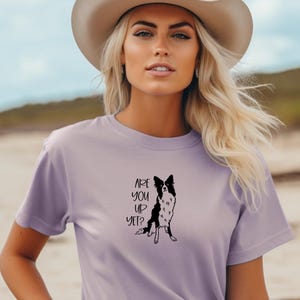 Puede incluir: Camiseta lila con un gr&aacute;fico en blanco y negro de un perro y el texto "ARE YOU UP YET?". La persona lleva un sombrero de vaquero beige. Prenda de vestir informal.