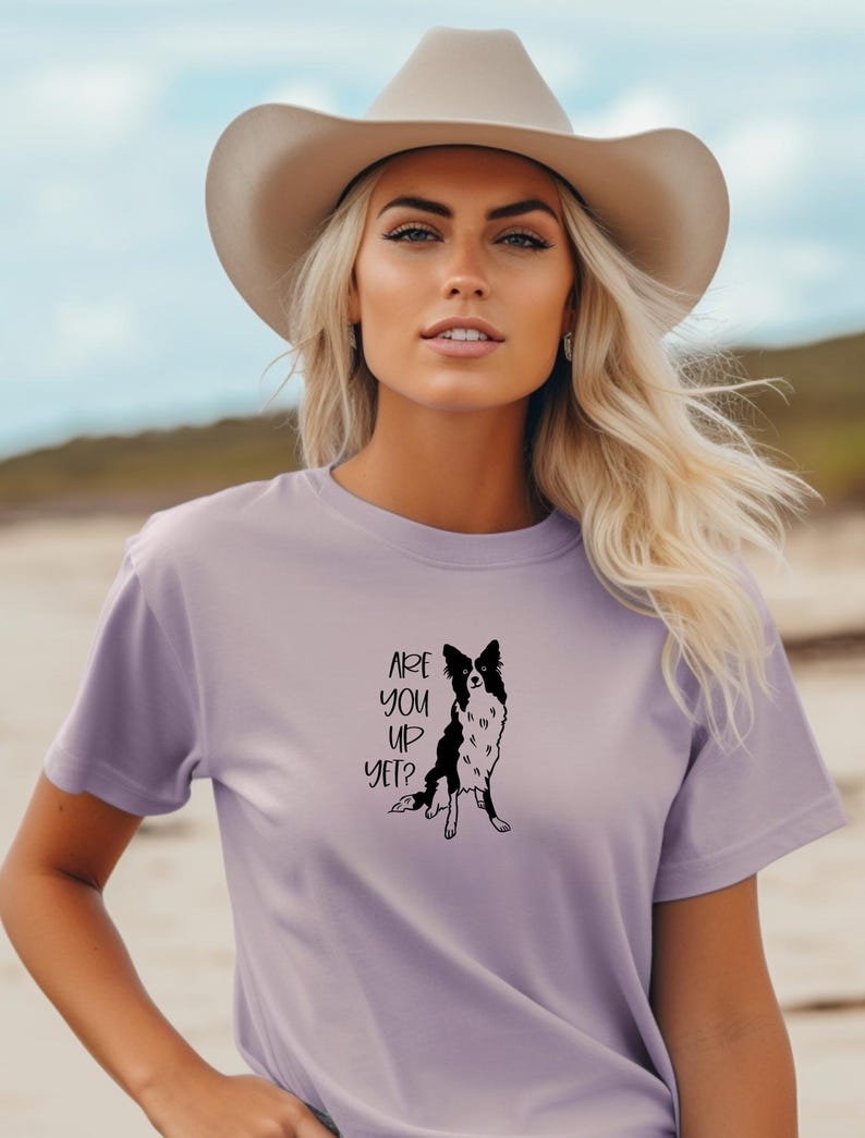 Puede incluir: Camiseta lila con un gr&aacute;fico en blanco y negro de un perro y el texto "ARE YOU UP YET?". La persona lleva un sombrero de vaquero beige. Prenda de vestir informal.