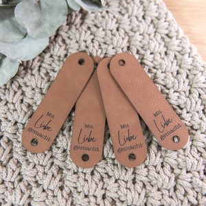 Könnte beinhalten: Vier braune Leder-Etiketten mit dem Text "Mit Liebe gemacht" in schwarzer Schrift.