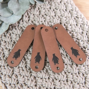 Könnte beinhalten: Vier braune Lederanhänger mit einem schwarzen Pinguin-Design. Jeder Anhänger hat oben ein Loch.