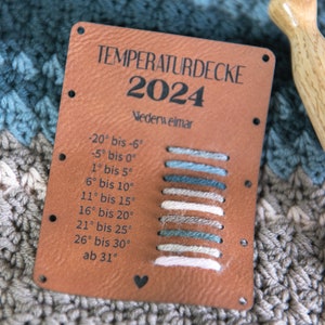 Könnte beinhalten: Ein braunes Leder-Etikett mit dem Text "TEMPERATURDECKE 2024 Nederweimar" und einer Temperaturtabelle mit den dazugehörigen Garnfarben. Die Tabelle zeigt Temperaturbereiche in Grad Celsius von -20 bis über 30 Grad.