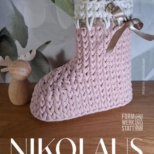 Puede incluir: Una bota de punto rosa con puño blanco y cinta de raso. La bota forma parte de un proyecto de manualidades, con la palabra "ANLEITUNG" en la parte superior y "NIKOLAUS Stiefel" en la inferior. A la izquierda hay una figura de madera.