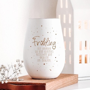 Könnte beinhalten: Ein weißer Kerzenhalter aus Keramik mit goldener Schrift "Frühling" und einem Zitat. Der Halter steht auf einem Holzsockel mit Lederriemen. Im Hintergrund befindet sich ein weißer, hausförmiger Kerzenhalter.