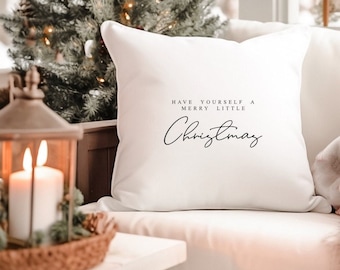 May include: White throw pillow with the text "Have yourself a merry little Christmas" in black script font. The pillow is sitting on a white sofa in front of a Christmas tree.