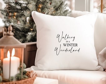 May include: White cushion with black text that reads "Walking in a Winter Wonderland". The cushion is on a white sofa with a Christmas tree in the background.