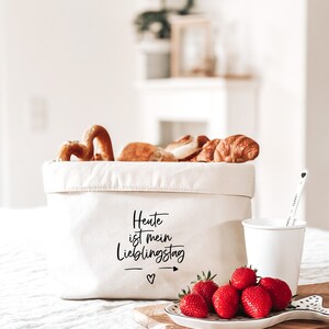 Op de afbeelding: Een witte canvas opbergmand met de tekst "Heute ist mein Lieblingstag" erop gedrukt. De mand is gevuld met brood en gebak. Voor de mand staat een wit keramiek bord met rode aardbeien op een houten snijplank.