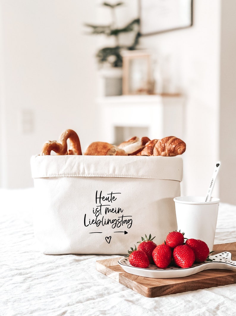 Op de afbeelding: Een witte canvas opbergmand met de tekst "Heute ist mein Lieblingstag" erop gedrukt. De mand is gevuld met brood en gebak. Voor de mand staat een wit keramiek bord met rode aardbeien op een houten snijplank.
