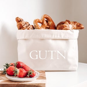 Op de afbeelding: Een witte canvas broodmand met het woord "GUTN" erop gedrukt. De mand is gevuld met croissants en pretzels. Een kleine witte kom met rode aardbeien staat op een houten snijplank.