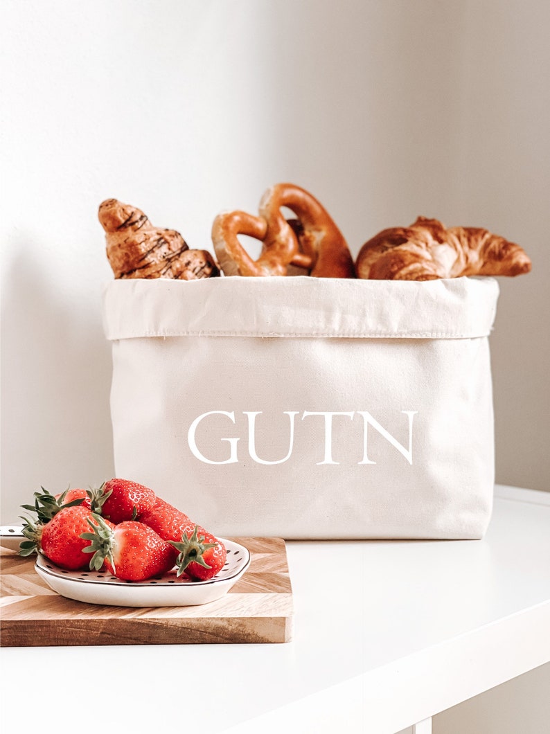 Op de afbeelding: Een witte canvas broodmand met het woord "GUTN" erop gedrukt. De mand is gevuld met croissants en pretzels. Een kleine witte kom met rode aardbeien staat op een houten snijplank.