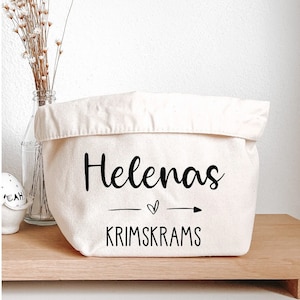 Könnte beinhalten: Eine weiße Stoffaufbewahrungstasche mit schwarzem Text, der "Helenas Krimkrams" mit einem kleinen Herz und einem Pfeil-Motiv lautet.
