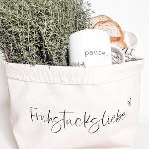 Op de afbeelding: Een witte canvas tas met de tekst "Fr&uuml;hst&uuml;ckliebe" en een klein hartje geprint op de voorkant. De tas is gevuld met verse tijm, een witte mok met het woord "pause" erop gedrukt en brood.