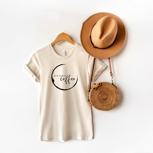 Könnte beinhalten: Ein beiges T-Shirt mit einem schwarzen Kaffeetassenring-Motiv und dem Text "But first, coffee".