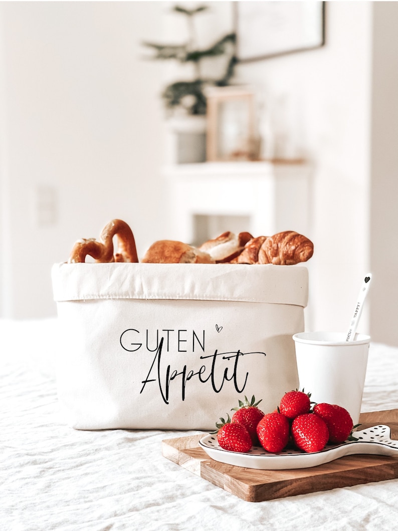 Op de afbeelding: Een witte canvas opbergbak met de tekst "Guten Appetit" in zwarte schrijfletters. De bak is gevuld met broodjes en croissants. Een witte mok en een bord met rode aardbeien staan op een houten snijplank.