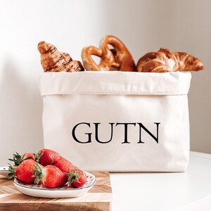Op de afbeelding: Een witte canvas broodmand met het woord "GUTN" erop gedrukt. De mand is gevuld met croissants en pretzels. Voor de mand staat een klein wit bord met rode aardbeien op een houten snijplank.