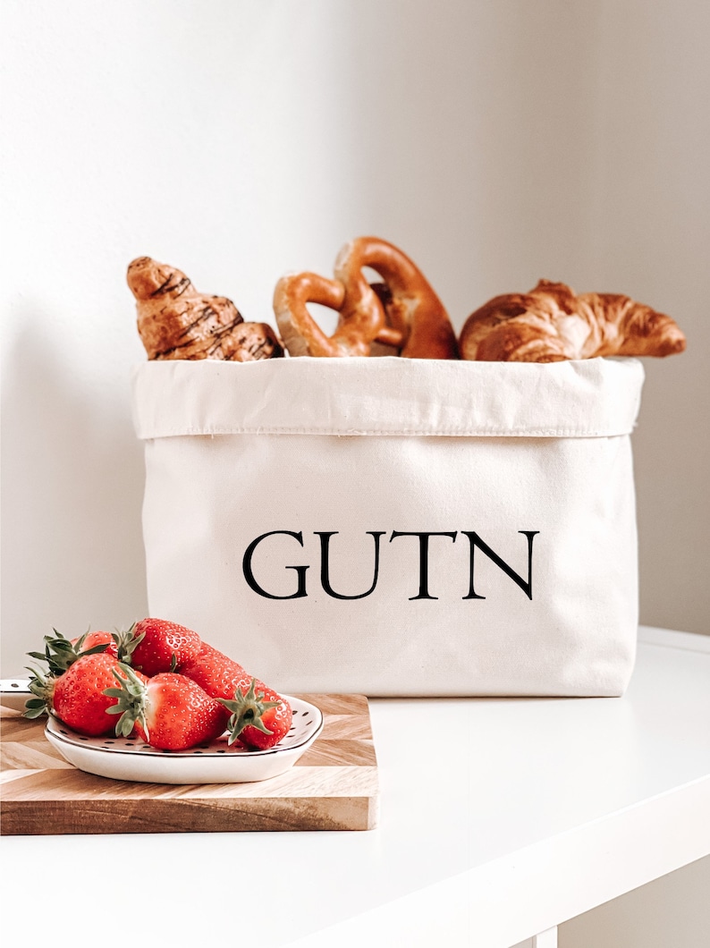 Op de afbeelding: Een witte canvas broodmand met het woord "GUTN" erop gedrukt. De mand is gevuld met croissants en pretzels. Voor de mand staat een klein wit bord met rode aardbeien op een houten snijplank.