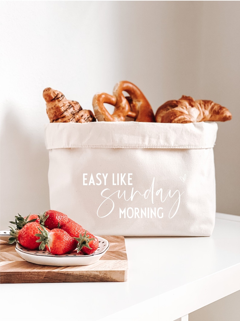 Op de afbeelding: Een witte canvas tas met de tekst "EASY LIKE sunday MORNING" erop gedrukt. De tas is gevuld met gebak, waaronder croissants en pretzels. Op de voorgrond staat een klein wit bord met rode aardbeien.