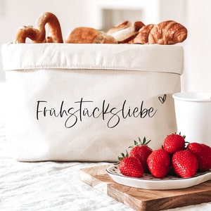 Op de afbeelding: Een witte canvasmand met de tekst "Fr&uuml;hst&uuml;ckliebe" en een klein hartje erop gedrukt. De mand is gevuld met broodjes en croissants. Een witte keramische mok en een witte keramische schaal met rode aardbeien staan op een houten oppervlak.