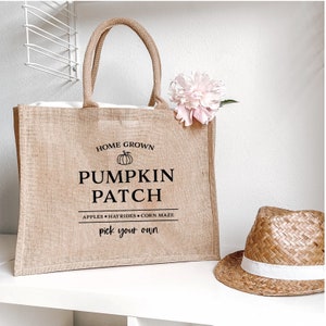 Könnte beinhalten: Eine braune Jute-Tragetasche mit schwarzem Text, der "Home Grown Pumpkin Patch Apples Hayrides Corn Maze Pick Your Own" lautet. Die Tasche hat ein weißes Futter und steht auf einer weißen Oberfläche. Ein Strohhut mit weißem Band liegt neben der Tasche.