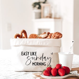 Op de afbeelding: Een witte canvas tas met de tekst "Easy Like Sunday Morning" in zwarte letters. De tas is gevuld met gebak, waaronder pretzels, croissants en kaneelbroodjes. Een witte keramische mok en een bord met rode aardbeien staan op een houten snijplank.