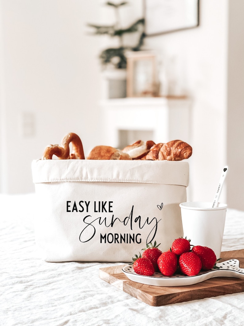 Op de afbeelding: Een witte canvas tas met de tekst "Easy Like Sunday Morning" in zwarte letters. De tas is gevuld met gebak, waaronder pretzels, croissants en kaneelbroodjes. Een witte keramische mok en een bord met rode aardbeien staan op een houten snijplank.
