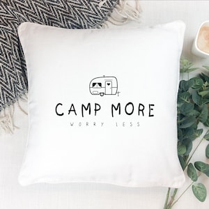 Könnte beinhalten: Weißes quadratisches Kissen mit einer schwarz-weißen Wohnwagenillustration und den Worten "CAMP MORE WORRY LESS" in Schwarz. Das Kissen liegt auf einer weißen Oberfläche, im Hintergrund eine schwarz-weiß gemusterte Decke.