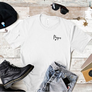 May include: White t-shirt with the word "Papa" and two small hearts printed on the front. The shirt is laid flat on a wooden surface with a pair of black boots, a beanie, sunglasses, and a key chain.