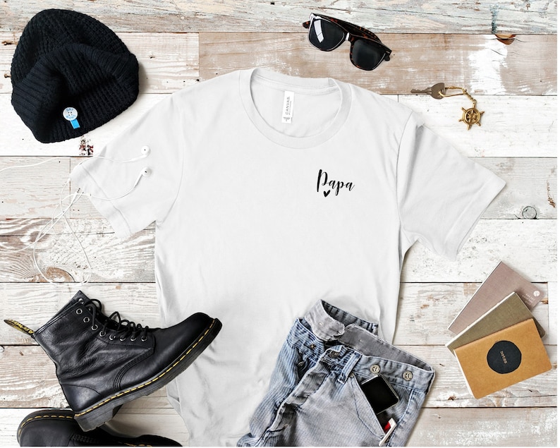 May include: White t-shirt with the word "Papa" and two small hearts printed on the front. The shirt is laid flat on a wooden surface with a pair of black boots, a beanie, sunglasses, and a key chain.