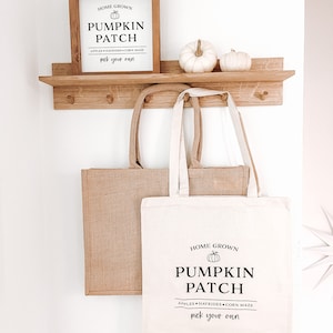 Könnte beinhalten: Ein Holzschild mit dem Text "Home Grown Pumpkin Patch Apples Hayrides Corn Maze Pick Your Own" und einer Schwarzweißillustration eines Kürbisses. Das Schild hängt an einem Holzregal mit zwei weißen Kürbissen auf dem Regal.