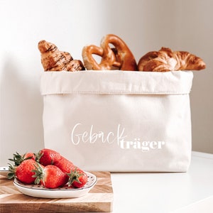 Op de afbeelding: Een witte canvas tas met de tekst "Geb&auml;cktr&auml;ger" erop gedrukt. De tas is gevuld met gebak, waaronder croissants en pretzels. Een klein wit schaaltje met rode aardbeien staat op een houten snijplank.