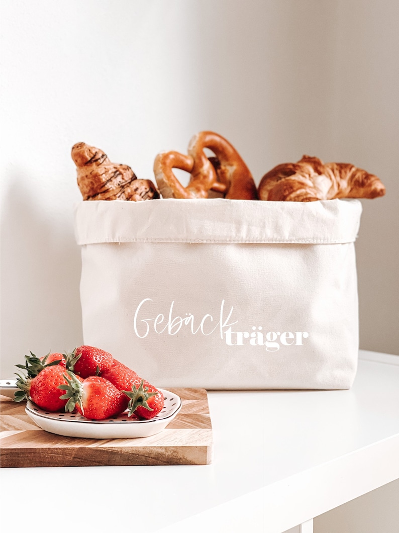 Op de afbeelding: Een witte canvas tas met de tekst "Geb&auml;cktr&auml;ger" erop gedrukt. De tas is gevuld met gebak, waaronder croissants en pretzels. Een klein wit schaaltje met rode aardbeien staat op een houten snijplank.