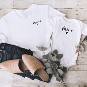 May include: White t-shirt with the word "mama" and a small heart printed on the front. A white onesie with the word "mini" and a small heart printed on the front. The shirts are laid out on a wooden surface with a pair of brown leather sandals, blue jeans, and a sprig of greenery.