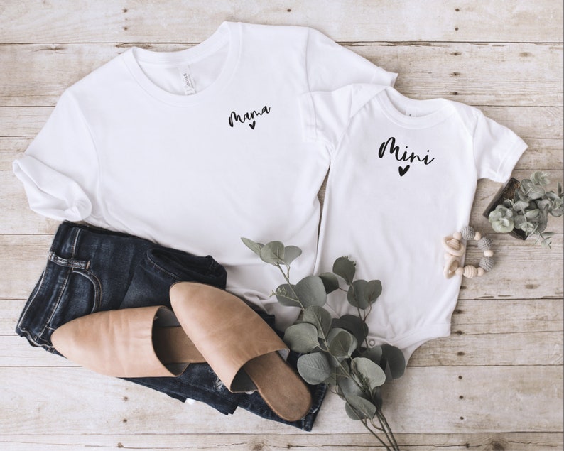 May include: White t-shirt with the word "mama" and a small heart printed on the front. A white onesie with the word "mini" and a small heart printed on the front. The shirts are laid out on a wooden surface with a pair of brown leather sandals, blue jeans, and a sprig of greenery.