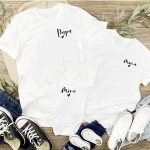 May include: Three white t-shirts with the words "Papa", "Mama", and "Mini" printed on them. Each shirt has a small heart symbol next to the text. The shirts are laid out on a wooden surface with blue jeans and white sneakers.