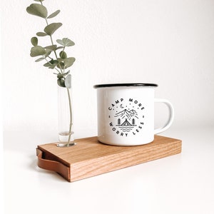 Könnte beinhalten: Eine weiße Emailletasse mit schwarzem Rand und Henkel. Die Tasse hat ein schwarz-weißes Grafikdesign mit dem Text "Camp More Worry Less" und einer Bergszene mit einem Zelt und einer Mondsichel. Die Tasse steht auf einem Holztablett mit einem Ledergriff. Neben der Tasse steht eine Glasvase mit einem Zweig Eukalyptus.