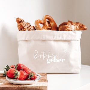 Op de afbeelding: Een witte canvas broodmand met de tekst "Br&ouml;tchen geber" en een klein hartje. De mand is gevuld met croissants en pretzels. Een kleine witte kom met rode aardbeien staat op een houten snijplank.