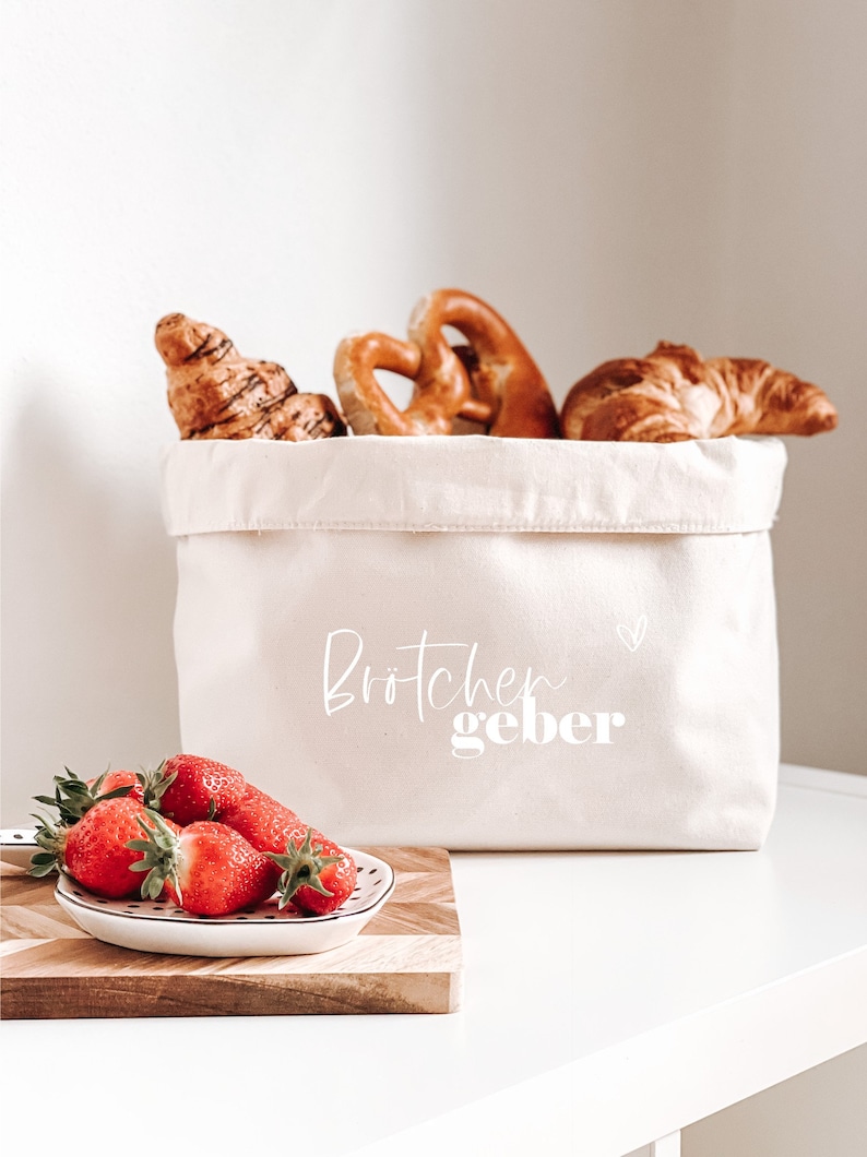 Op de afbeelding: Een witte canvas broodmand met de tekst "Br&ouml;tchen geber" en een klein hartje. De mand is gevuld met croissants en pretzels. Een kleine witte kom met rode aardbeien staat op een houten snijplank.