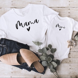 Könnte beinhalten: Weißes T-Shirt mit dem Text "mama" in schwarzer Schrift mit einem kleinen Herz darunter. Ein weißer Baby-Strampler mit dem Text "mini" in schwarzer Schrift mit einem kleinen Herz darunter.