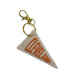 Puede incluir: Llavero con banderín triangular con la palabra "CLEVELAND" bordada en hilo naranja. El banderín es de color blanquecino con un cierre y un anillo de metal dorado.