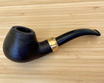 May include: A black wooden pipe with a gold band and a black mouthpiece. The pipe is resting on a wooden surface.